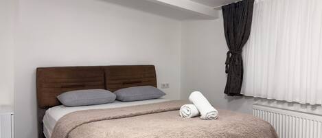 A bedroom with a bed, pillows, a rolled towel, a window with curtains, and a radiator.