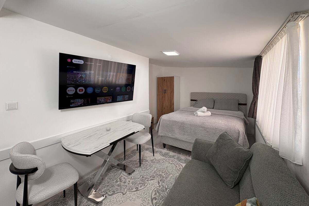 A modern hotel room with a large flat-screen TV mounted on the wall, a bed with grey bedding, a grey sofa, and a small desk with a chair.