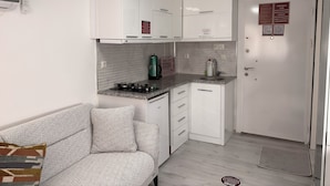 Une cuisine de taille réduite, avec des meubles blancs, une cuisinière à gaz, une machine à café et un réfrigérateur.