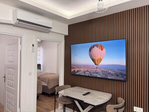 A modern room with a mounted TV displaying a hot air balloon, a dining table with chairs, a bed, and an air conditioning unit. - Saklıkonak Otel (Isparta)