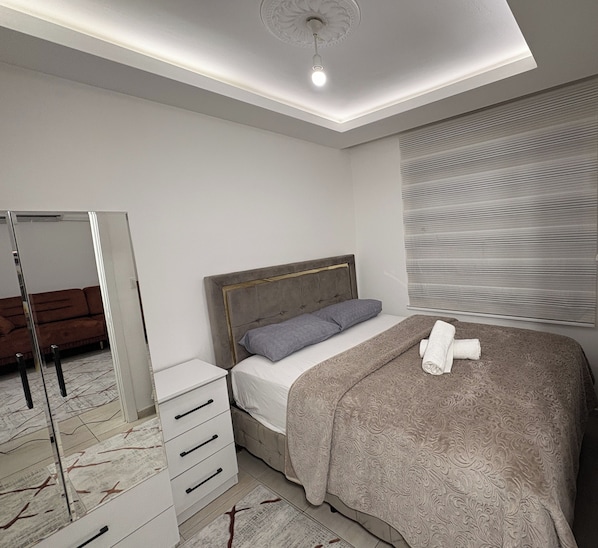 A bedroom with a bed, bedside table, dresser, mirror, and a ceiling light. - Saklıkonak Otel (Isparta)