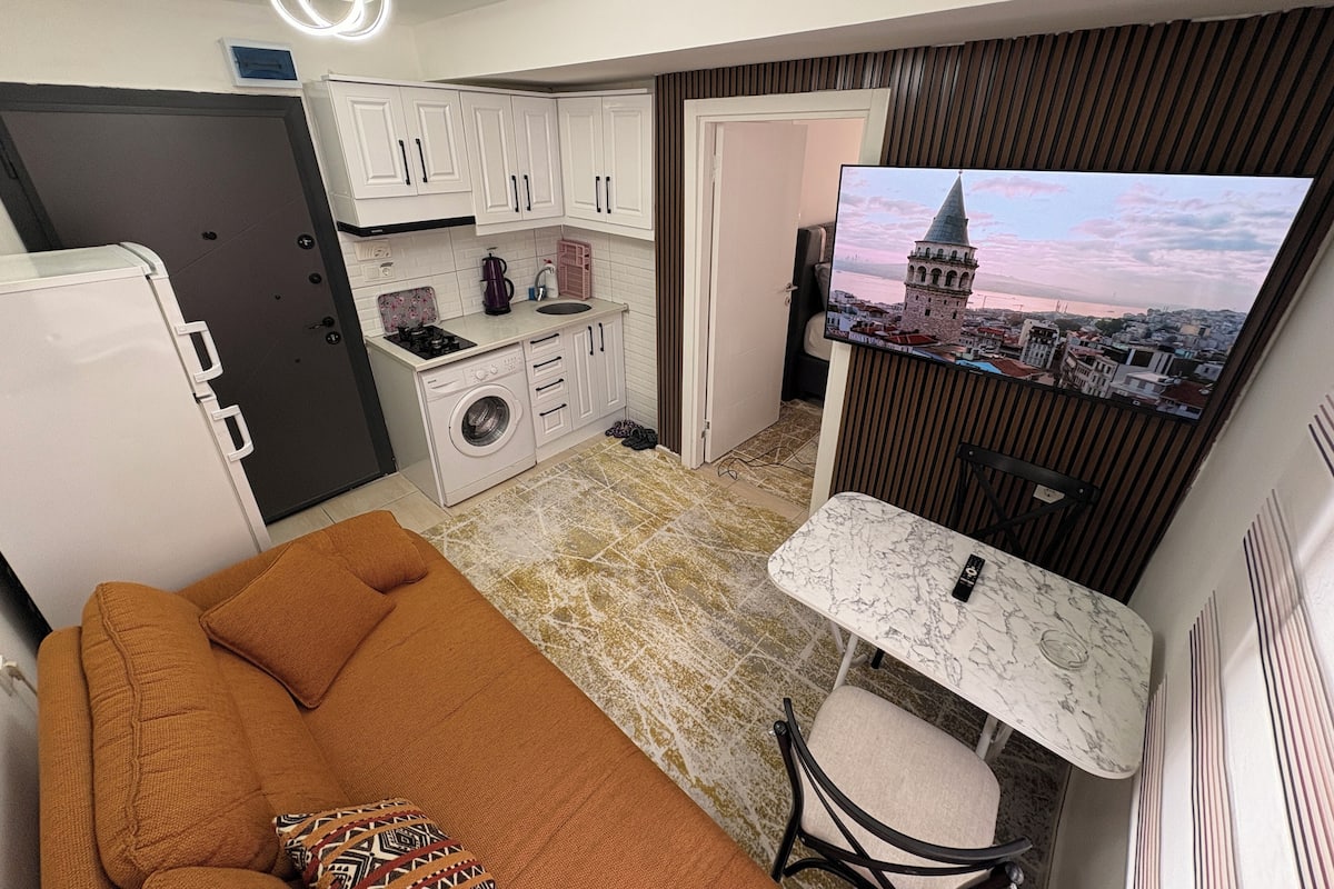 A compact living space with a kitchenette, a sofa, a small table, and a TV displaying a cityscape.