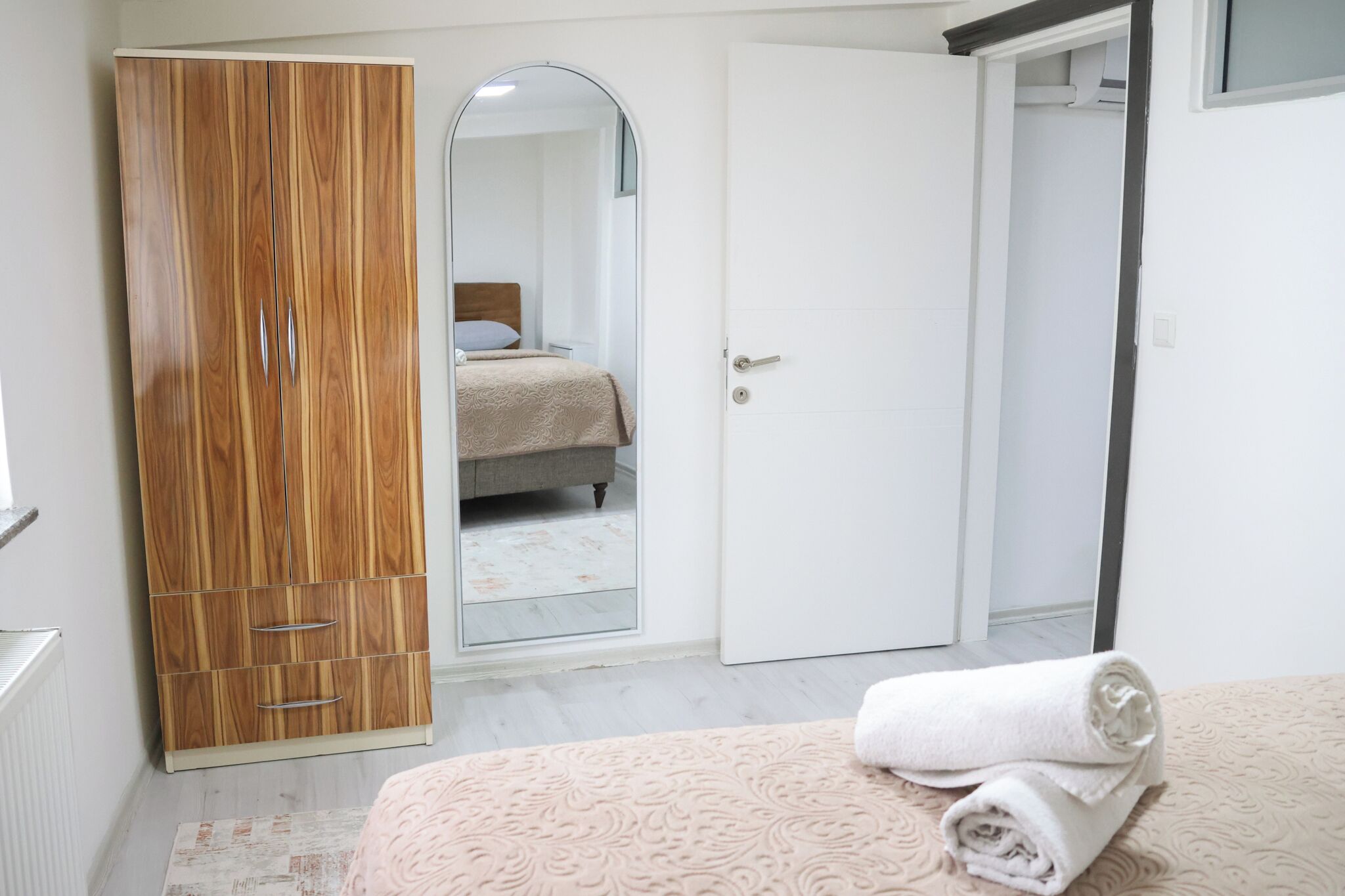 A bedroom with a wooden wardrobe, a bed with a rolled towel, and a door leading to another room.