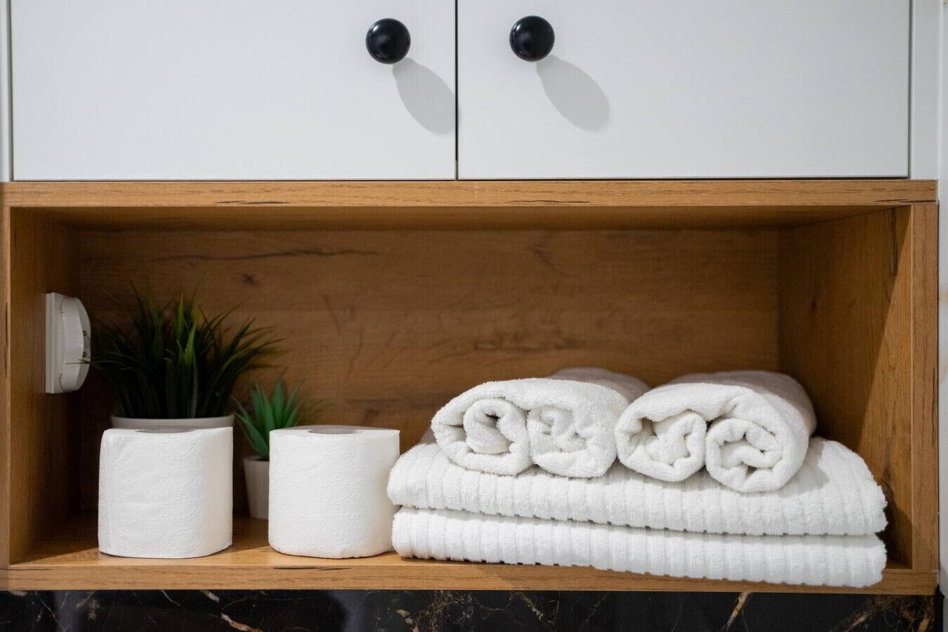 Une salle de bain avec une étagère en bois sur laquelle sont posés des serviettes enroulées, un rouleau de papier toilette et une plante en pot.