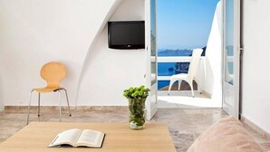A room with a view of the sea, a flat-screen TV mounted on the wall, a wooden table with an open book, a chair, and a potted plant. - 1809 Villa (Santorini)
