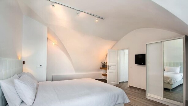 A modern bedroom with a large bed, a walk-in closet, and a television on the wall. - Thireas Hotel (Santorini)