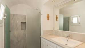 A bathroom with a glass-enclosed shower, a white sink, and a large mirror. - Dimitrios Cave House (Santorini)