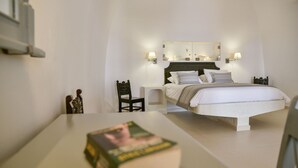 A bedroom with a large bed, a nightstand, a chair, and a bedside table. - Dimitrios Cave House (Santorini)
