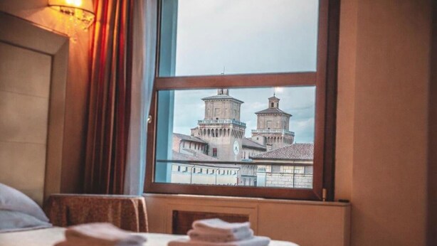 A hotel room with a bed, a window with a view of historic buildings, and a bedside table with towels. - The Dome - Panoramic Rooms (Ferrara)