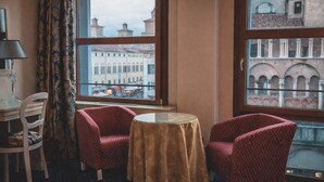A hotel room with two red patterned chairs, a small round table, a lamp, and a view of a cityscape through large windows. - The Dome - Panoramic Rooms (Ferrara)