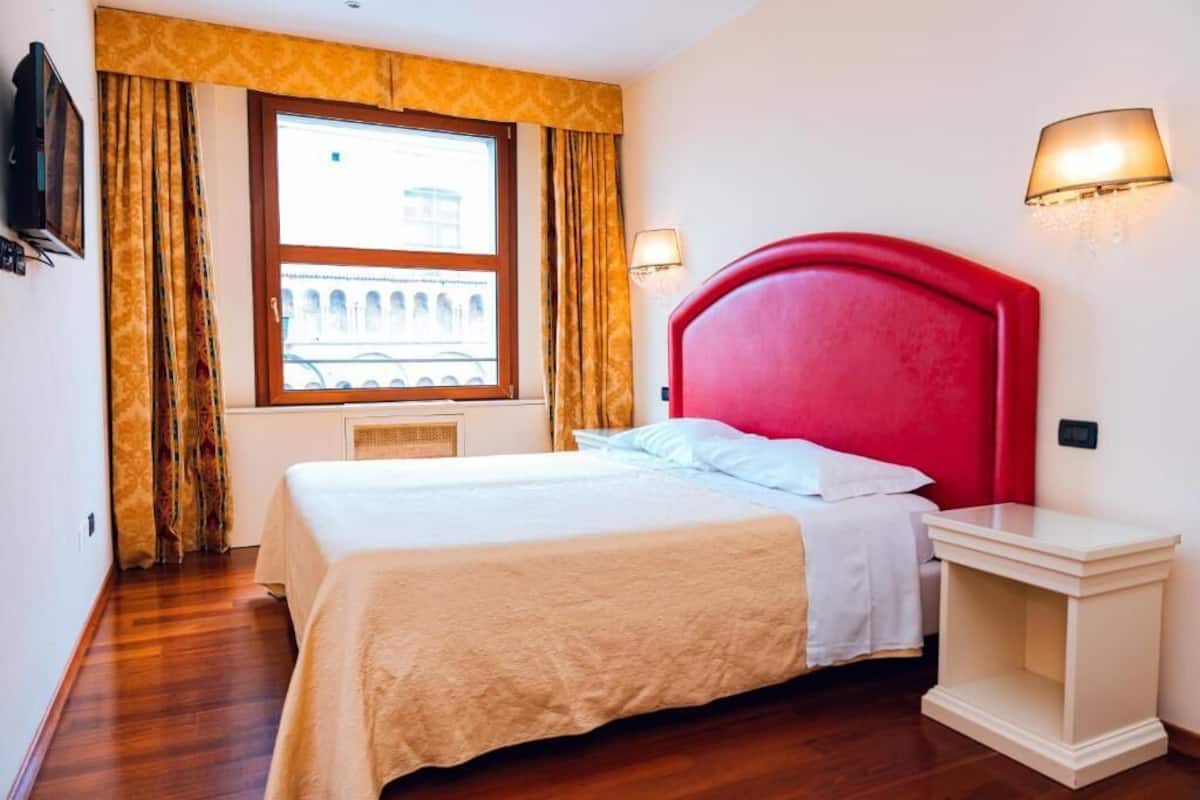 A hotel room with a large bed, a window with a view of a building, a red headboard, and two wall-mounted lamps.