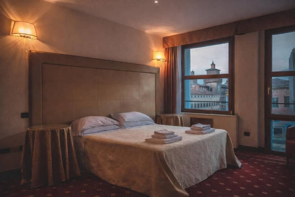 A hotel room with a large bed, a view of a cityscape through a window, and a red carpeted floor.