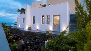 A modern white building with multiple windows and a door, surrounded by greenery and a rocky landscape. - San Marino Suites (Santorini)