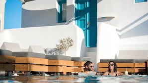 A poolside area with wooden loungers, a white building with blue accents, and a clear blue sky. - San Marino Suites (Santorini)