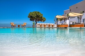 A pool area with lounge chairs and umbrellas, a tree, and a building in the background. - San Marino Suites (Santorini)