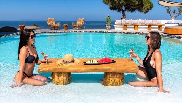 Two women sitting by a poolside table with drinks and food, overlooking a clear blue sea and a tree-lined deck. - San Marino Suites (Santorini)