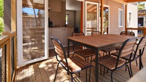 A wooden deck with a table and chairs, a sliding glass door leading to a kitchen, and a pergola overhead. - Bungalow in Sitges, Barcelona for 4-people (Sitges)