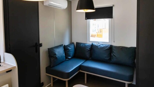 A modern room with a sectional sofa, two pendant lights, an air conditioning unit, and a window with a view. - Bungalow in Sitges, Barcelona for 4-people (Sitges)