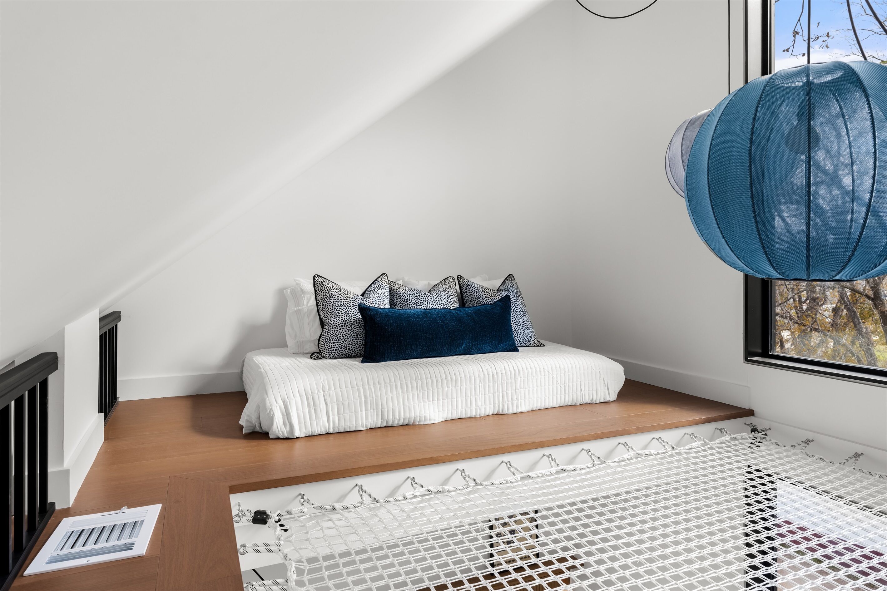 Een knus loft-slaapkamer met een wit bed, gekleurde kussens, een blauwe plaid en een hangende blauwe lamp.