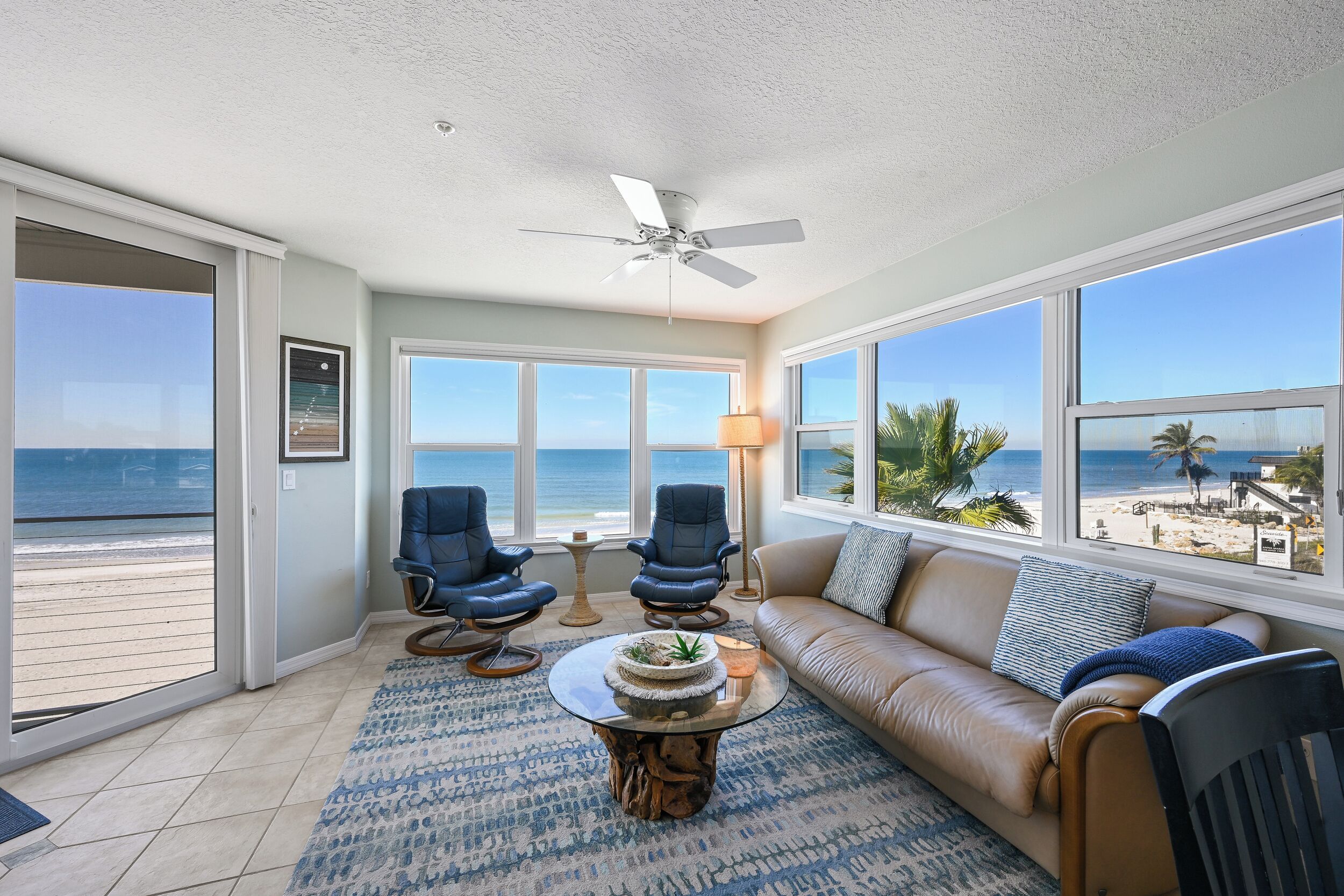 A spacious living room with a large window overlooking the beach, a ceiling fan, and a leather sofa.