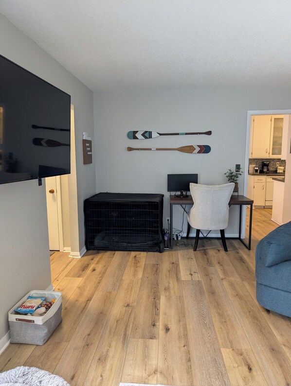 A living room with a black pet crate, a desk with a computer, and a TV mounted on the wall.