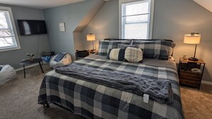 Une chambre à coucher avec une couvre-lit à carreaux, deux lampes de chevet, un téléviseur fixé au mur et une fenêtre avec des stores.