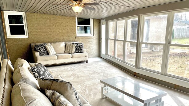 Ein geräumiges Wohnzimmer mit einem beige farbenen Sofa, gemusterten Kissen, einem Glastisch und großen Fenstern mit Blick auf den Garten.