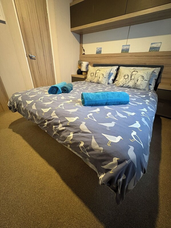 A neatly made bed with a patterned bedspread, two rolled towels, and matching pillows on a headboard with artwork. - 'Serenity' A Tranquil & Peaceful Retreat set in a Cornish country park (Relubbus)