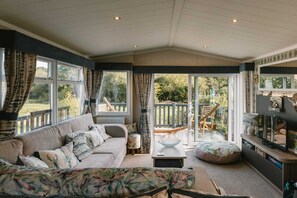 A cozy living room with a sofa, a coffee table, and a TV stand. There is a sliding glass door leading to an outdoor deck. - 'Serenity' A Tranquil & Peaceful Retreat set in a Cornish country park (Relubbus)