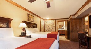 Ein Hotelzimmer mit zwei Betten, einem Schreibtisch, einem Stuhl, einem Deckenventilator und einem durchgegangenen Badezimmer.