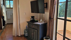 A room with a tiled floor, a flat-screen TV mounted on the wall, a cabinet with a coffee maker, a water dispenser, and a trash bin.