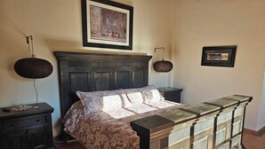 A bedroom with a large bed, dark wooden headboard, two bedside tables, a nightstand, a large round lamp, and a framed painting on the wall.