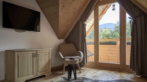 Een kamer met een houten plafond, een aan de muur bevestigde televisie, een houten kast, een stoel, een groot raam met gordijnen, en uitzicht op een houten terras.