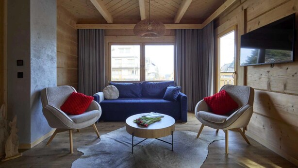 Een gezellige woonkamer met een blauwe bank, twee fauteuils, een ronde salontafel en een houten plafond.
