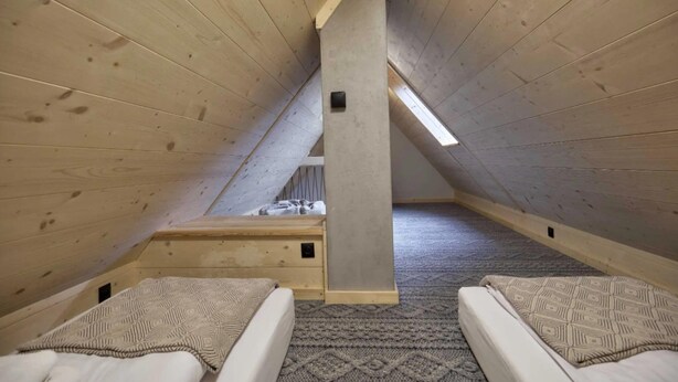 A small attic bedroom with two beds, a window, and a wooden ceiling.