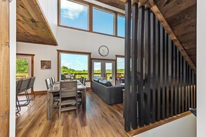 A spacious dining area with a long wooden table and chairs, a black leather sofa, and a large window with a view of greenery. - Amazing 5BD Pool Lodge | Theater + Hot Tub + Games (Sevierville)