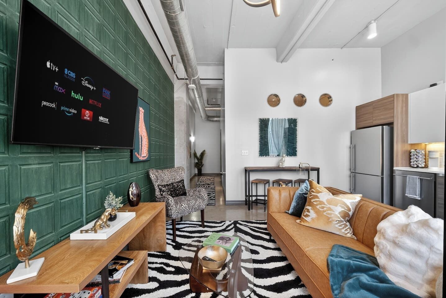 A modern living room with a flat-screen TV displaying streaming services, a zebra-patterned rug, a wooden coffee table, a brown leather sofa, and a green accent wall.