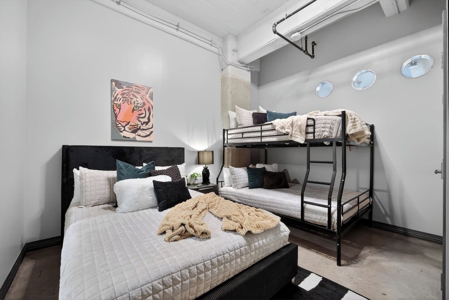 A dorm-style room with bunk beds, a large bed with multiple pillows, a tiger painting on the wall, and a small table with a lamp.