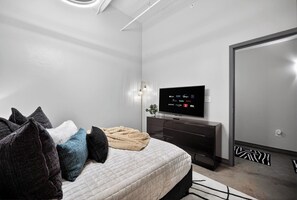 Un dormitorio moderno con cama, televisor mostrando servicios de streaming, cómoda y espejo.
