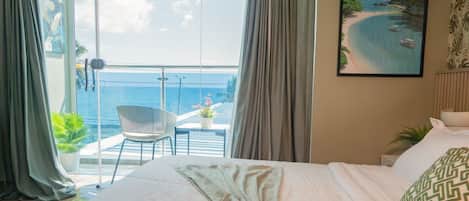 A hotel room with a large bed, a view of the ocean, a balcony with a chair, and a painting of a beach scene.