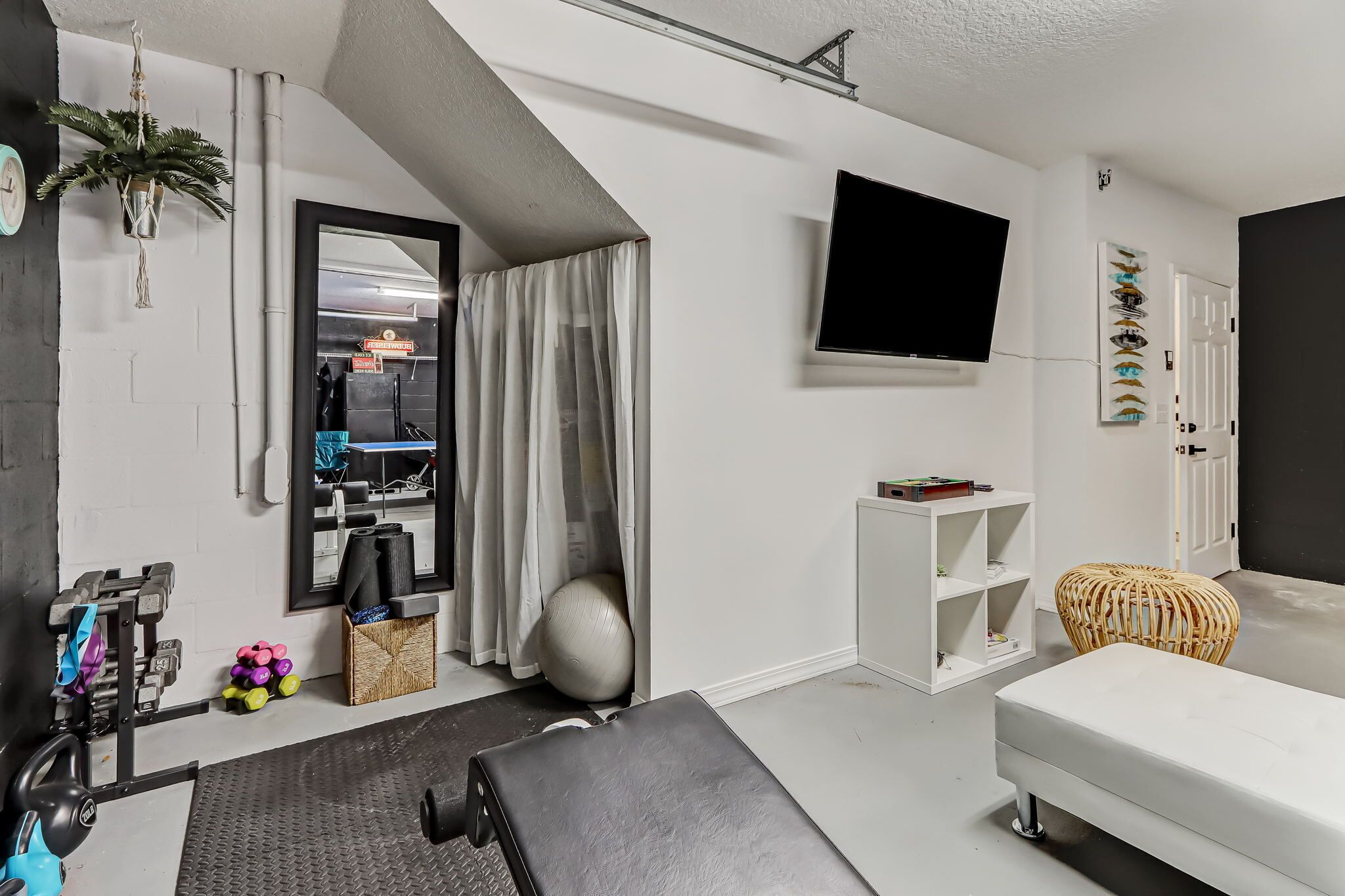 Una sala moderna con un sofá blanco, un televisor montado en la pared, una zona de entrenamiento con mancuernas y una colchoneta de yoga, y una pelota de ejercicios grande.
