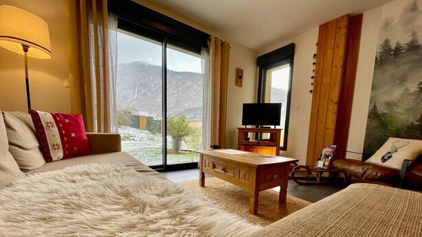 A bedroom with a large bed, a wooden nightstand, a television, and a view of the outdoors. - Gite Le 1000 Votre gîte à Loudenvielle - Comfort, nature and serenity (LOUDENVIELLE)