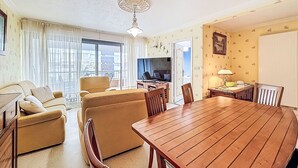 A living room with a sofa, chairs, a wooden dining table, a TV, and a balcony with curtains. - Kérabus by Interhome (Quiberon)