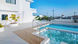 Ein moderner Poolbereich mit blauem Himmel, einem Pool mit Whirlpool und einem weißen Gebäude mit großen Fenstern und Pflanzen in Töpfen.