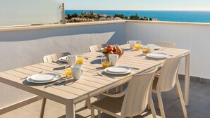 Ein Esstisch mit Tellern, Besteck, Gläsern und einer Obstschale auf einem Balkon mit Blick auf das Meer.