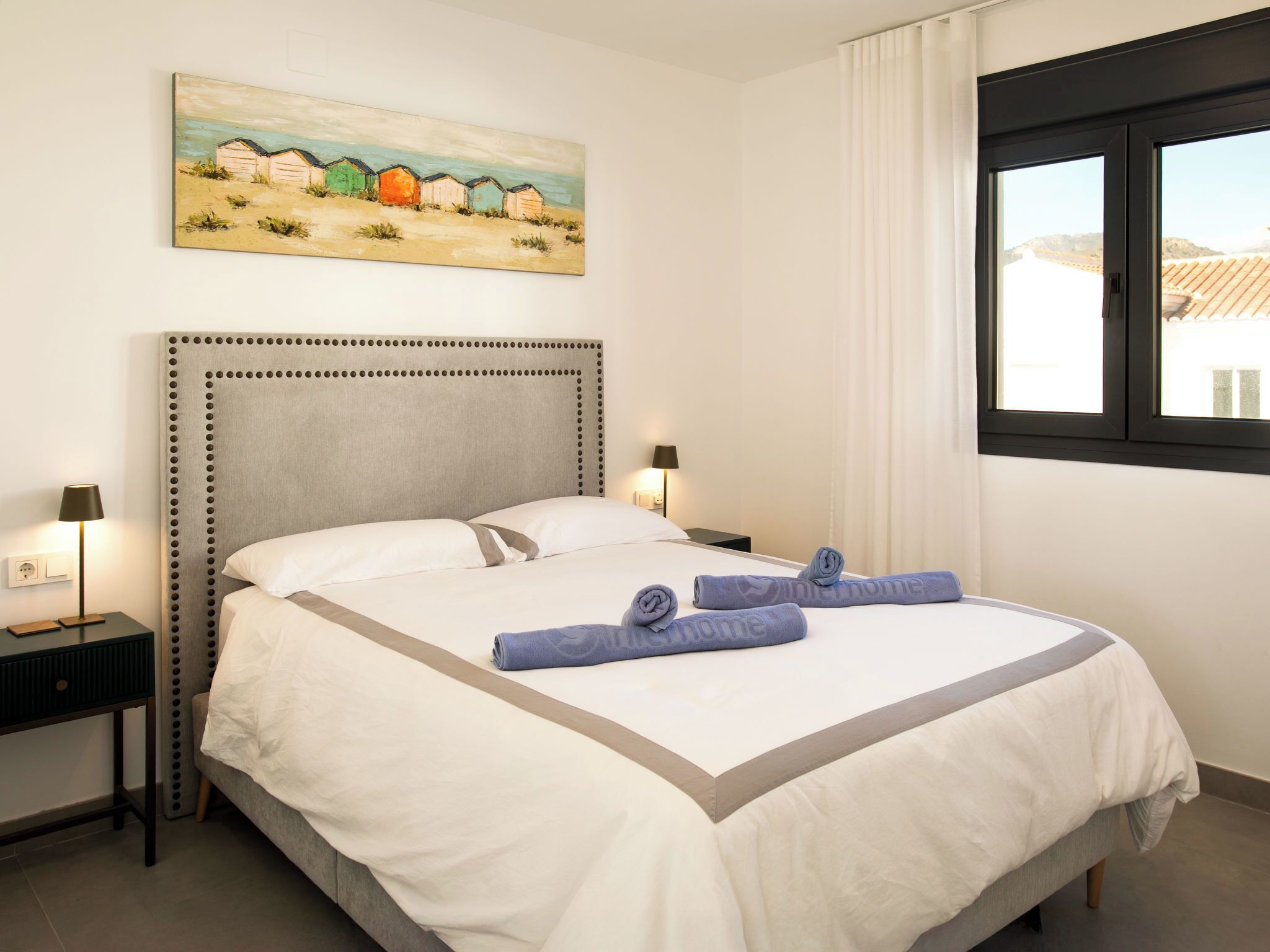 A neatly made bed with a headboard, two bedside tables with lamps, a painting of beach huts, and a window with curtains.