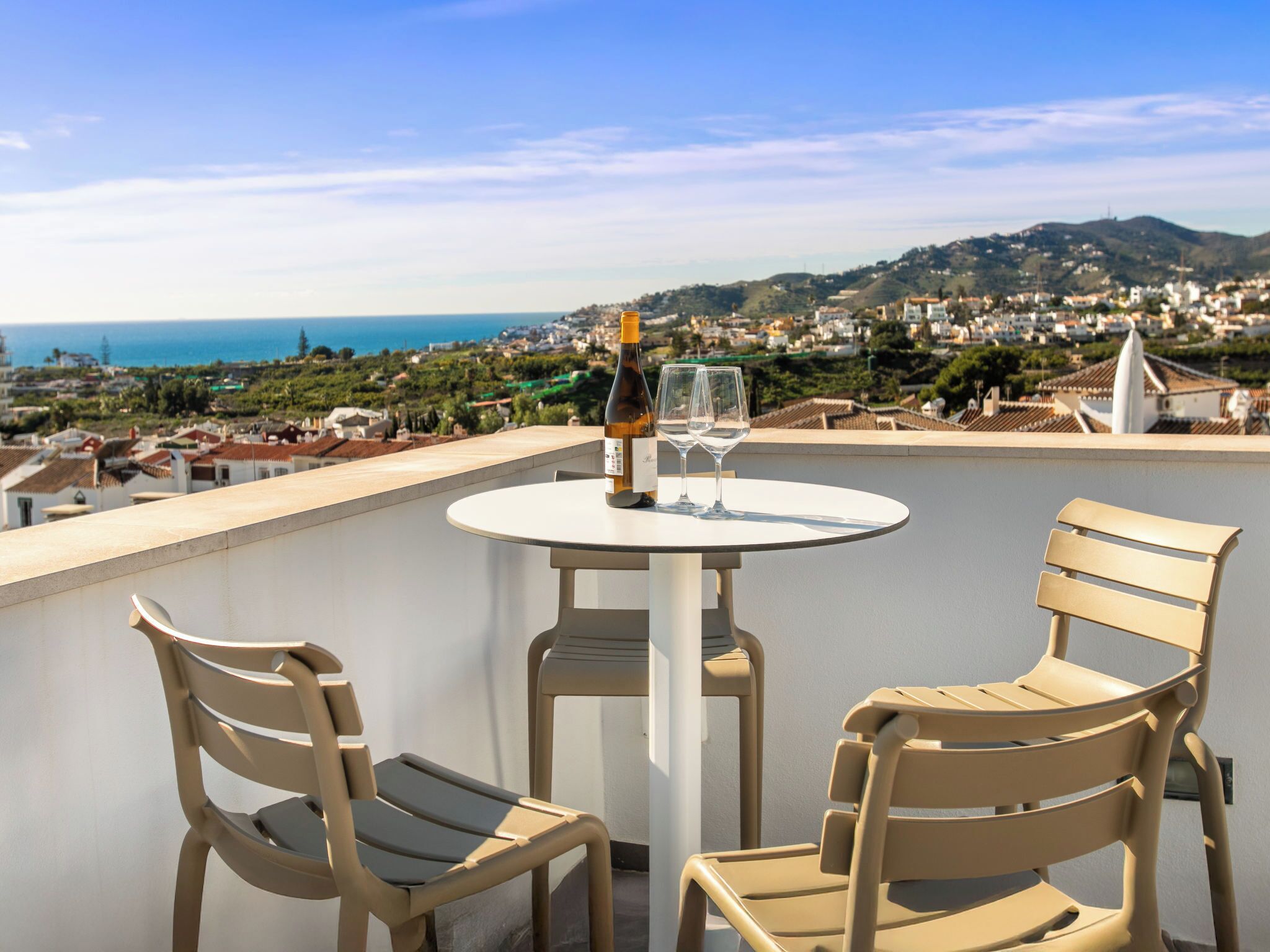 A terrace with a table and two chairs, overlooking a coastal town with hills and buildings.