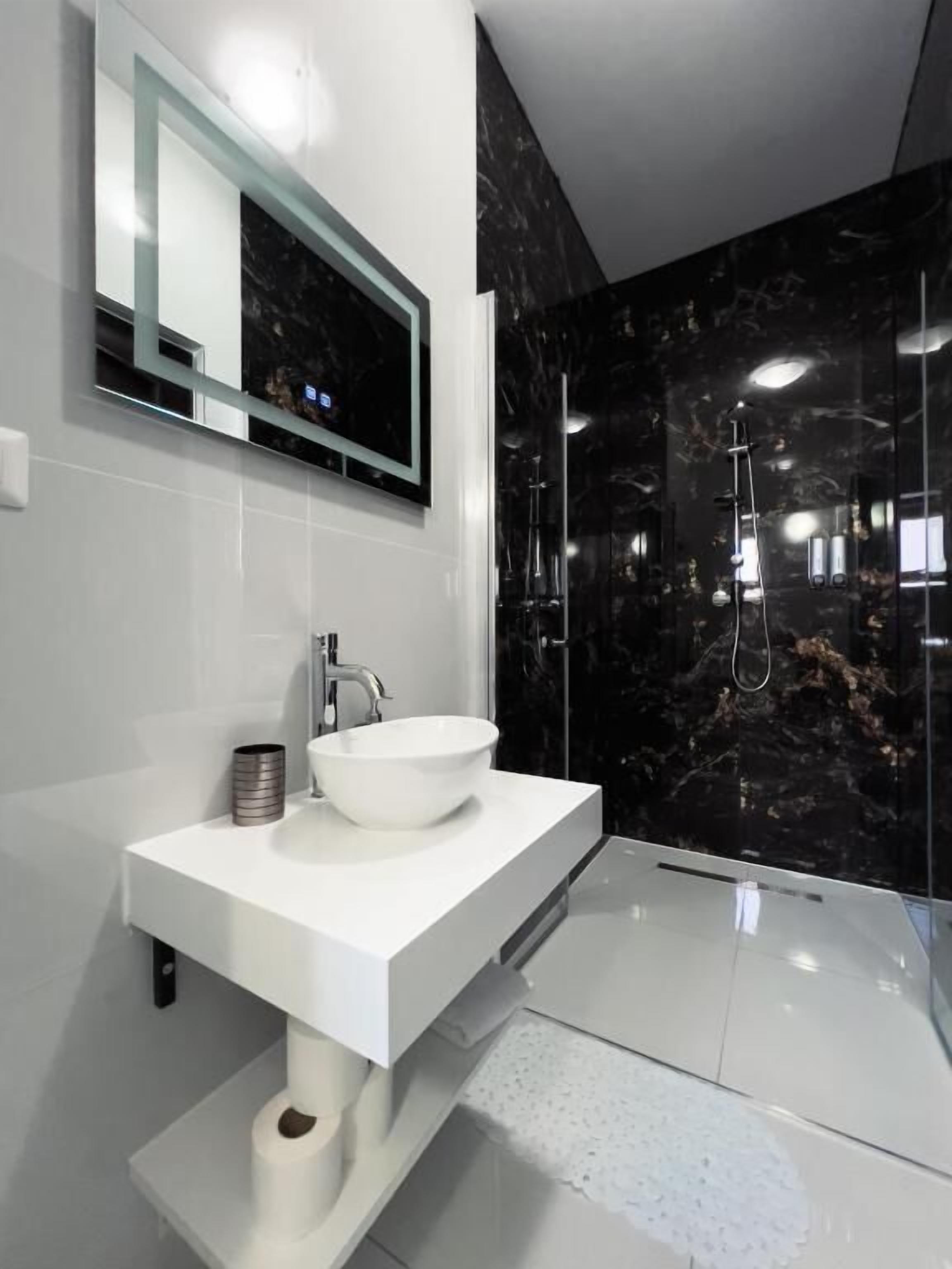 Un baño moderno con un tocador blanco, un espejo grande y un revestimiento de mármol negro en la pared de la ducha.