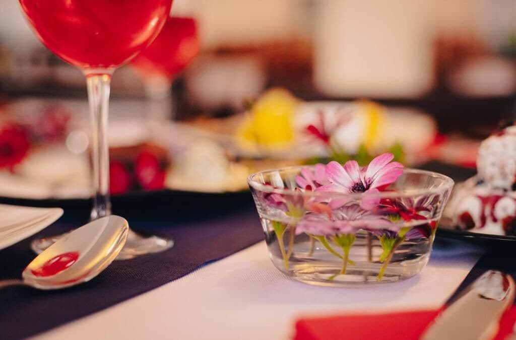 A dining table with a glass bowl containing pink flowers, a spoon, and a glass of red liquid.