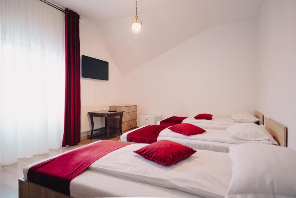 Quarto de hotel com três camas, cada uma com lençóis brancos e almofadas vermelhas. Há uma secretária de madeira, uma cadeira e uma televisão de tela plana na parede.